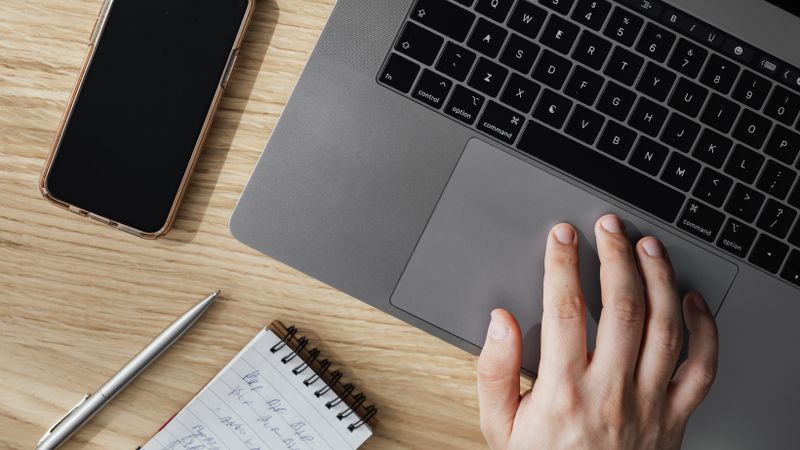 Mano sobre el trackpad de una notebook, al lado de un celular y una libreta con anotaciones